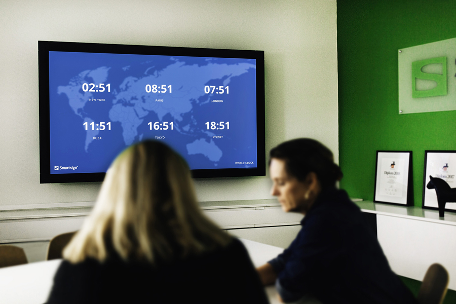 Digital signage-skärm i mötesrum som visar världsklocka med tider för olika städer som New York, Paris, Tokyo och Sydney, vilket illustrerar global kommunikation och informationsdelning i modern arbetsmiljö