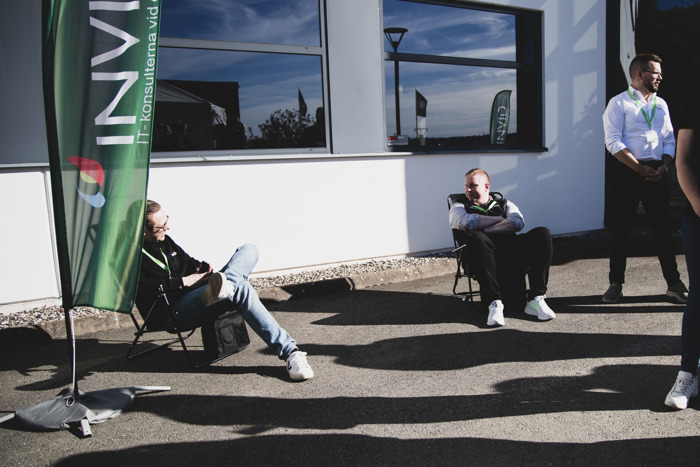 Deltagare sitter och står utomhus i solen under event, vid en byggnad med INVID‑flagga i bakgrunden.