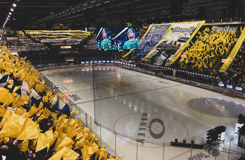 HV71-match i fullsatt arena med gul flaggtifo, publik och isplan, fokus på hockeyevent och stark lagstämning