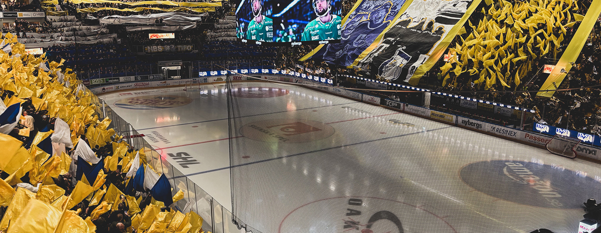 HV71-match i fullsatt arena med gul flaggtifo, publik och isplan, fokus på hockeyevent och stark lagstämning