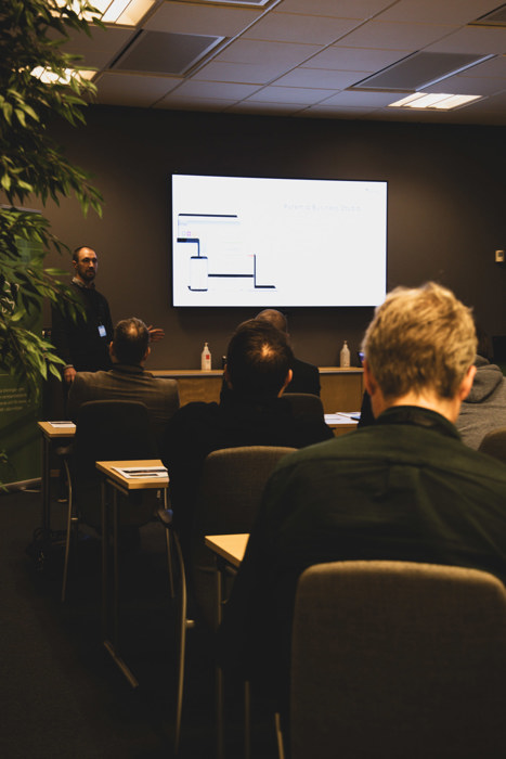 Seminarium i ett konferensrum där Linus Berglund presenterar vid en skärm med diagram, medan deltagare sitter vid bord med anteckningar och lyssnar