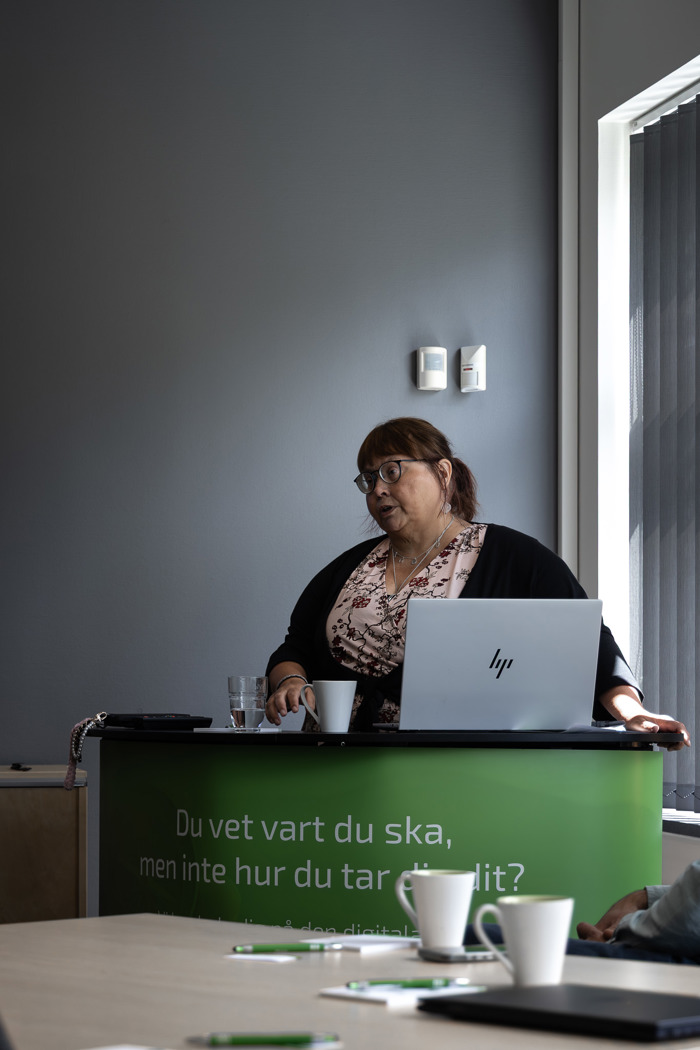 Anne-Marie Achrenius vid podium med laptop håller presentation i mötesrum, med kaffe och anteckningar på bordet i förgrunden