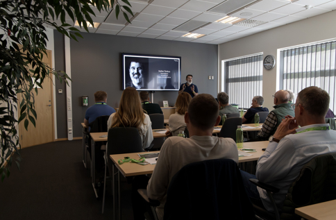Deltagare sitter i ett konferensrum och lyssnar på en presentation vid en stor skärm med porträttbild och text, medan en talare står längst fram och presenterar