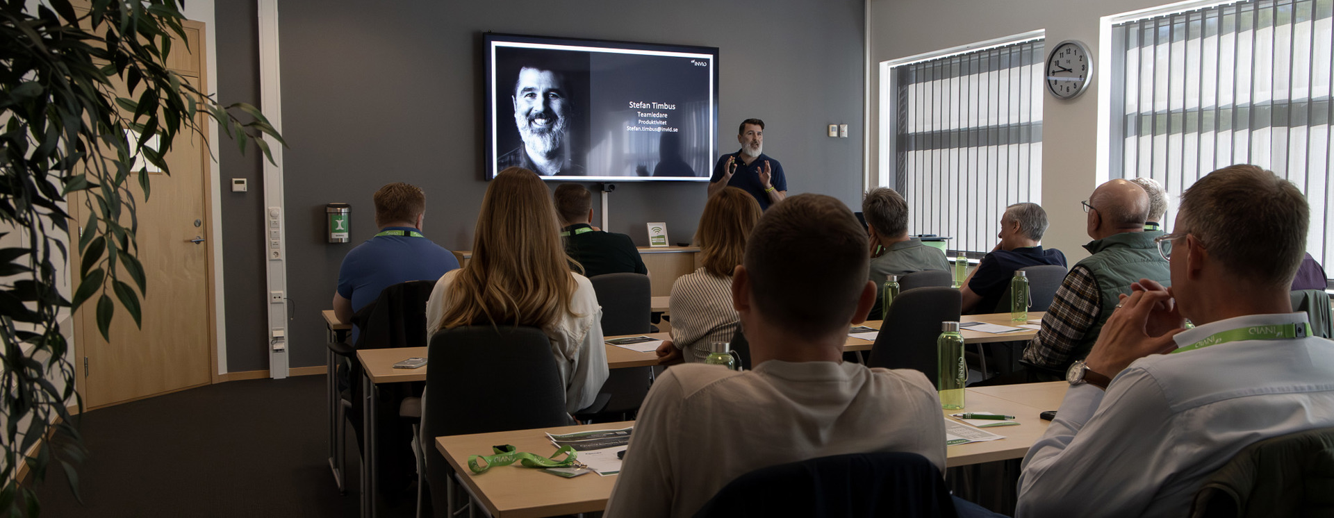 Deltagare sitter i ett konferensrum och lyssnar på en presentation vid en stor skärm med porträttbild och text, medan en talare står längst fram och presenterar