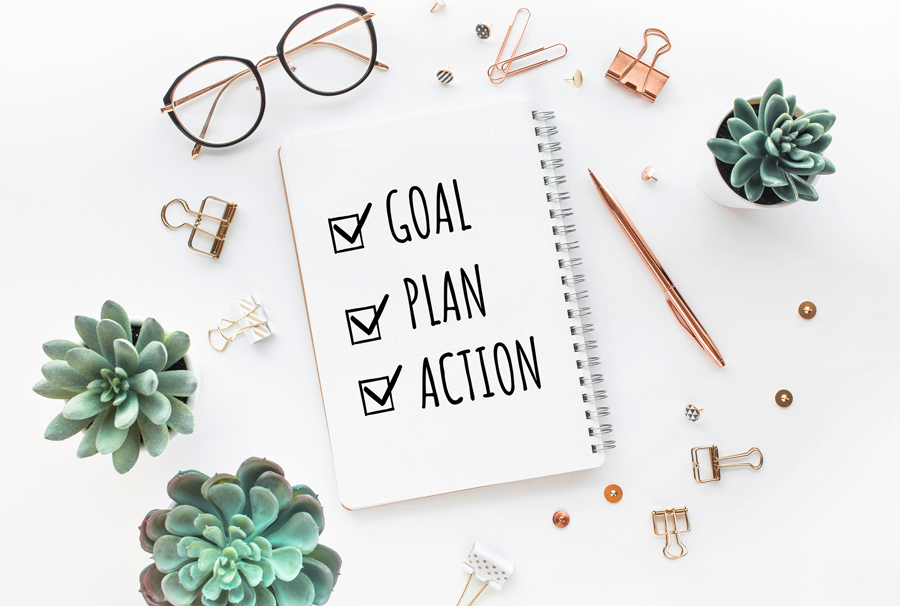 Anteckningsbok med checklista “Goal, Plan, Action”, omgiven av växter, glasögon och kontorsmaterial på ett skrivbord