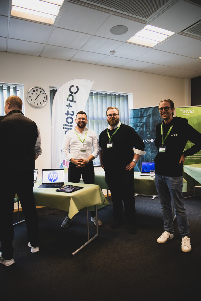 Flera personer står vid en demostation med laptops under event, med roll‑up för Copilot+ PC och teknikvisning i bakgrunden.