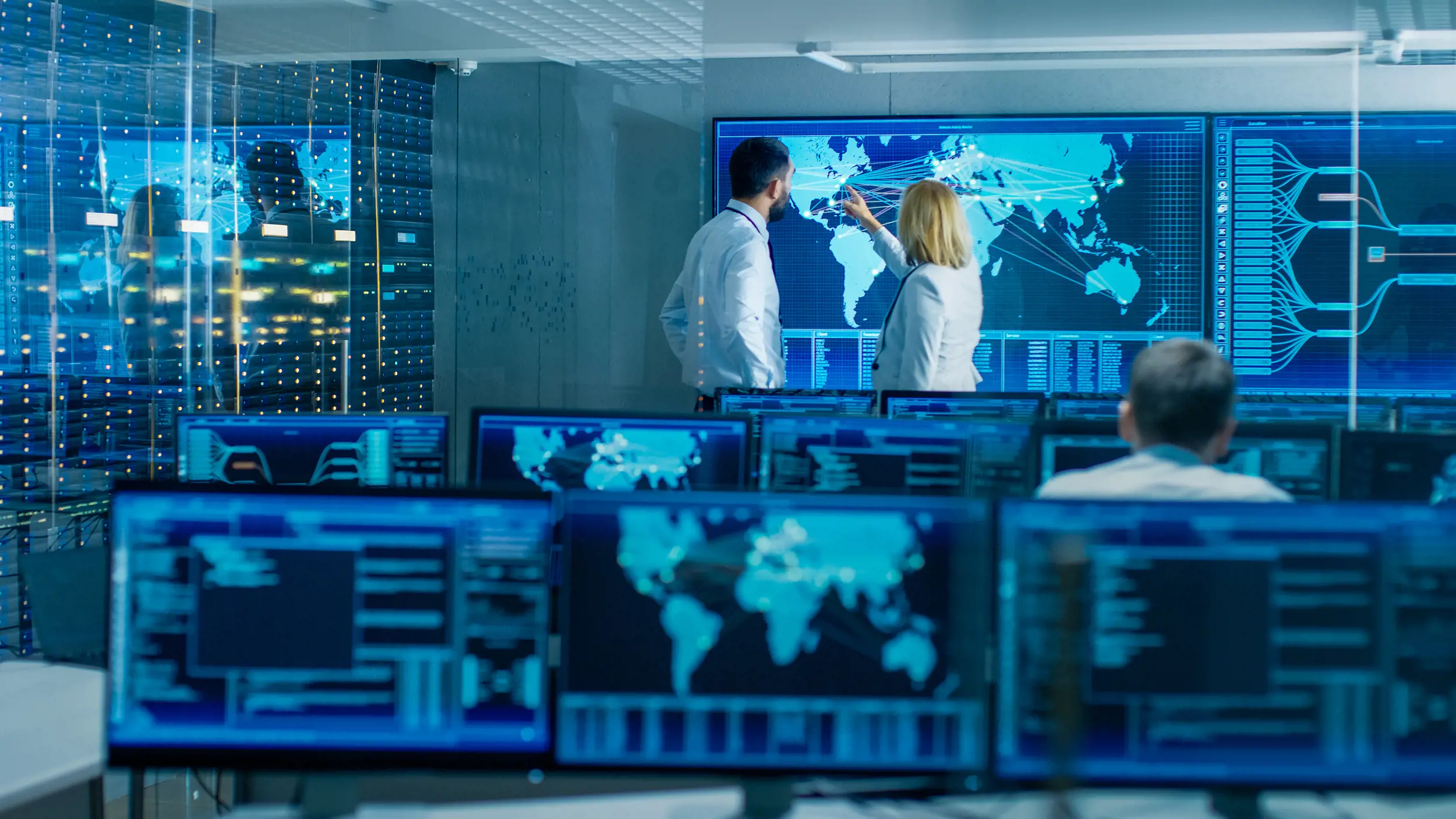 Flera personer arbetar i ett kontrollrum med skärmar som visar världskartor, nätverk och systemdata, där en person pekar på en global visualisering – vilket symboliserar omvärldsbevakning, hotanalys och IT‑övervakning