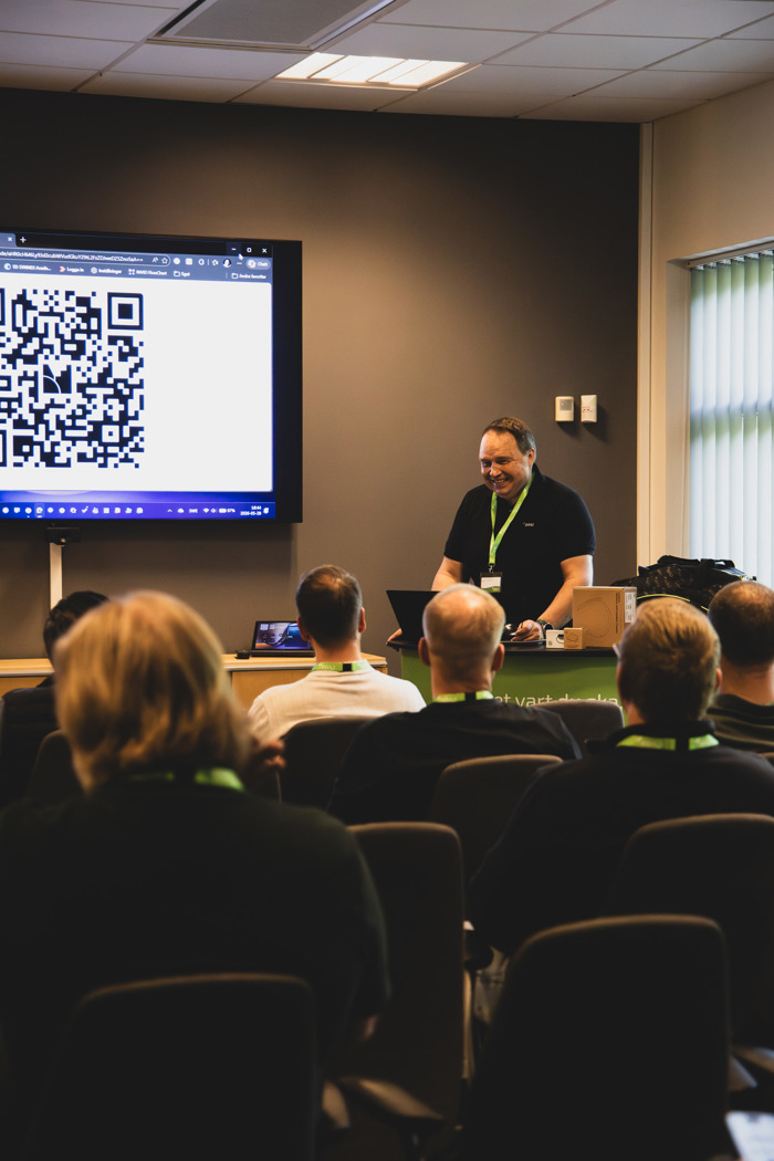 Föreläsare presenterar inför sittande publik i konferensrum, med QR‑kod visad på skärm bredvid presentationen.