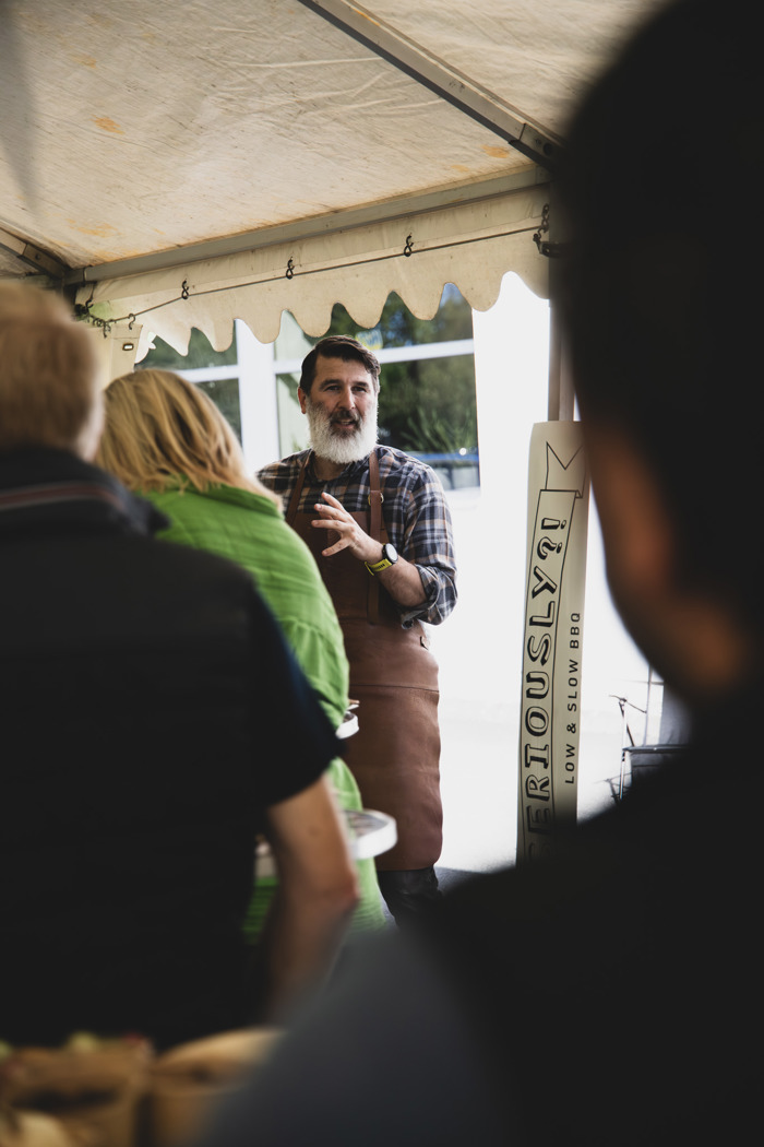 Person i förkläde presenterar och pratar med en grupp deltagare under utomhusevent i tält, med grill- eller matstation i bakgrunden.