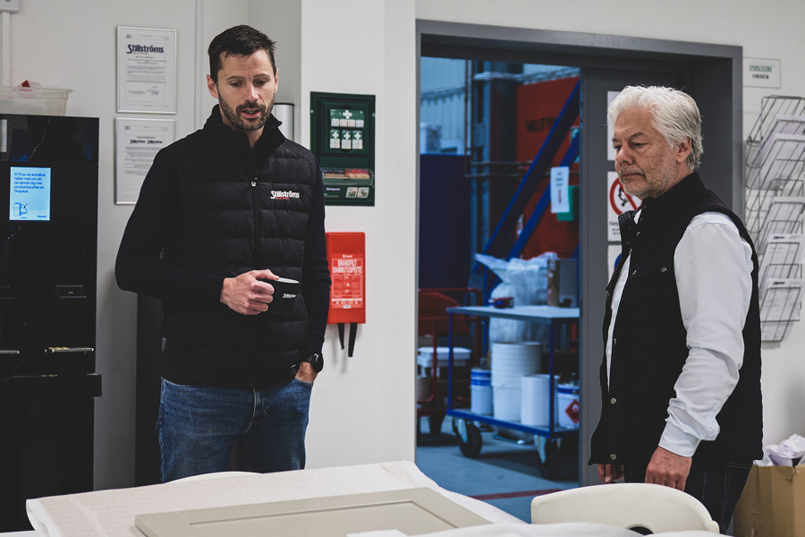 Två personer diskuterar vid ett arbetsbord i en industrimiljö med produktionslokal i bakgrunden
