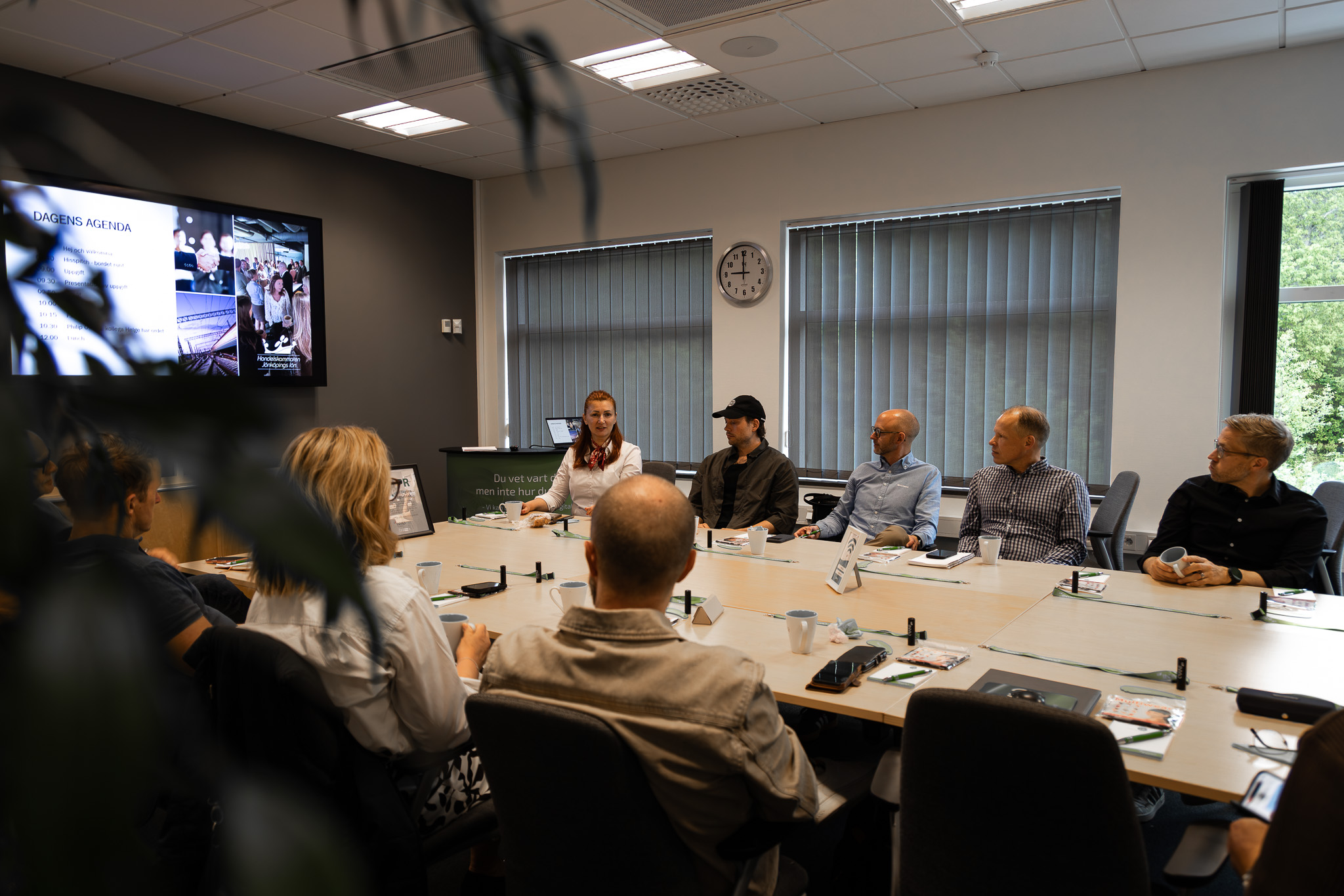 Personer sitter runt ett konferensbord i ett modernt mötesrum med laptoppar, anteckningsmaterial och kaffekoppar, under en presentation som visas på en skärm på väggen