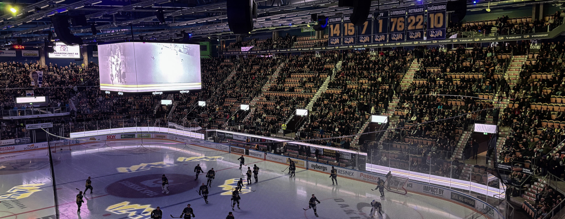 Hockeymatch i fullsatt arena med spelare på isen, storbildsskärm, publik på läktarna och exponering av INVID:s varumärke