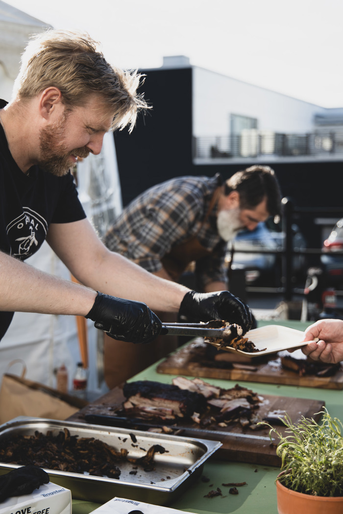 Person serverar grillat kött från skärbräda till tallrik vid utomhusgrill under event, med matförberedelser och tält i bakgrunden.