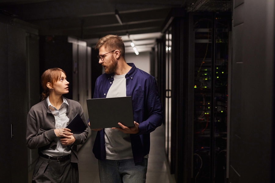 Två personer går genom serverhall med laptop och surfplatta och diskuterar drift och övervakning av IT‑infrastruktur, vilket illustrerar serverdrift som tjänst, samarbete och hantering av affärskritiska system i datacentermiljö