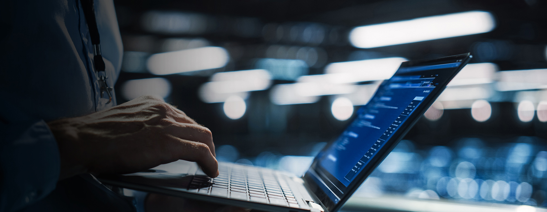 Person arbetar på laptop i serverhall med bakgrund av datacenter och nätverksutrustning, vilket visar övervakning, drift och underhåll av IT‑miljö och affärssystem i realtid