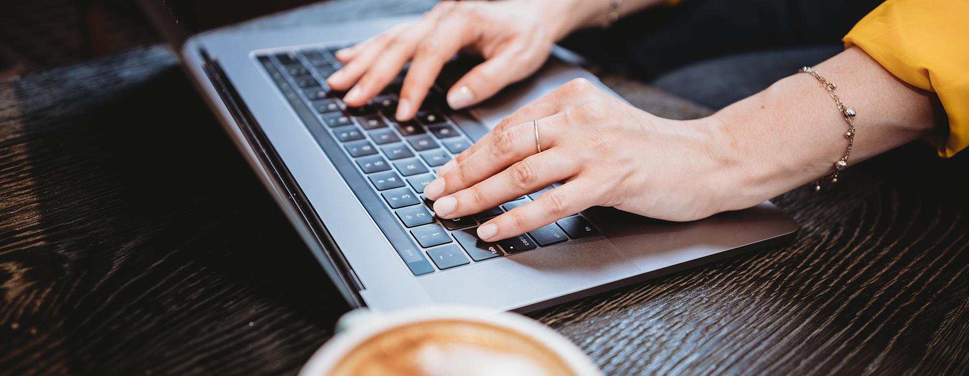 Närbild av händer som skriver på en laptop på ett bord, med en kopp kaffe i förgrunden
