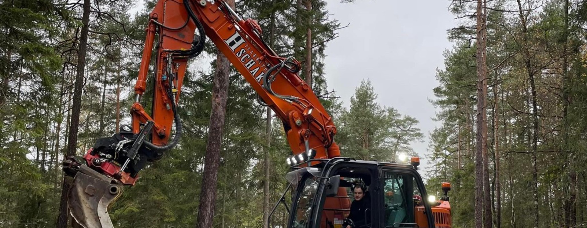 Bandgrävare arbetar med mark- och anläggningsarbete i skogsmiljö där väg eller infrastruktur byggs med hjälp av tung entreprenadutrustning