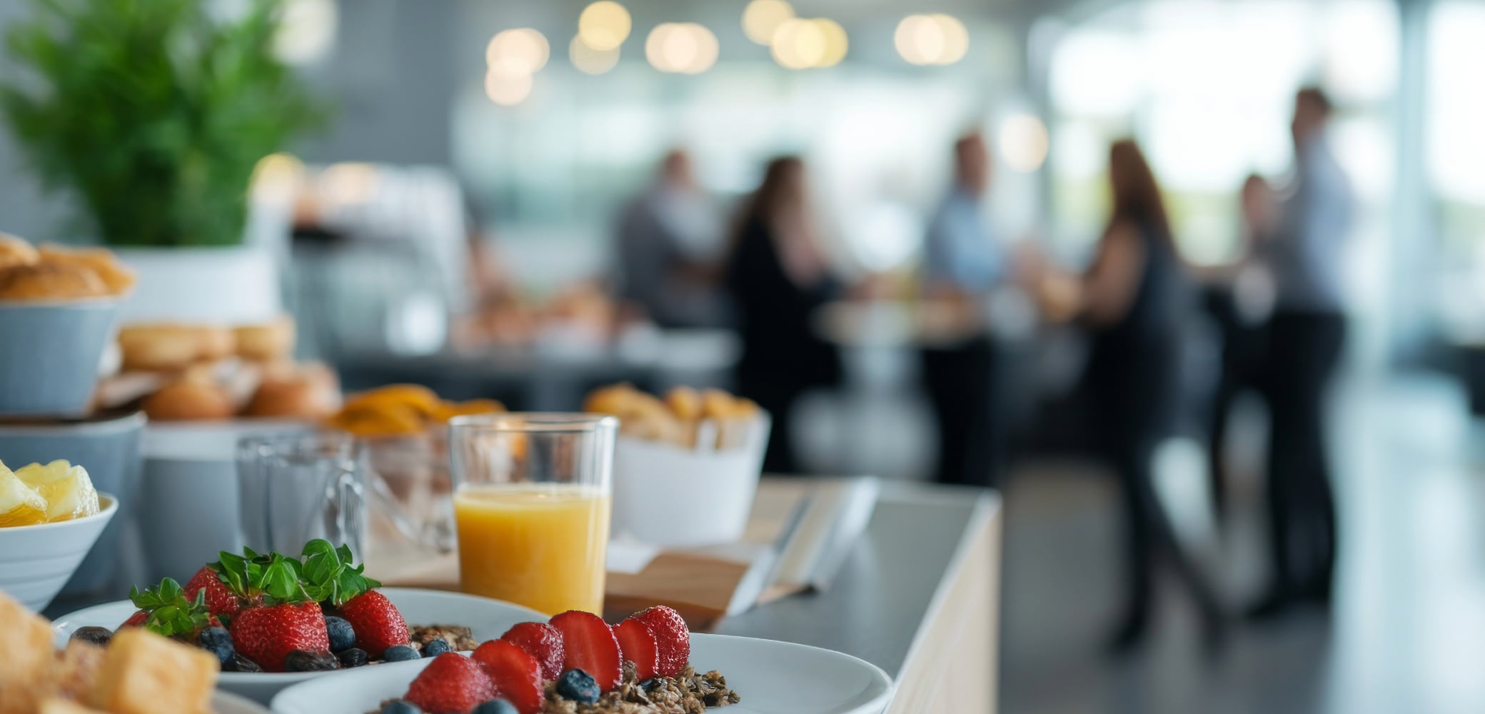 Frukostbuffé med jordgubbar, bröd och juice i förgrunden, människor samtalar suddigt i bakgrunden i ljus lokal.