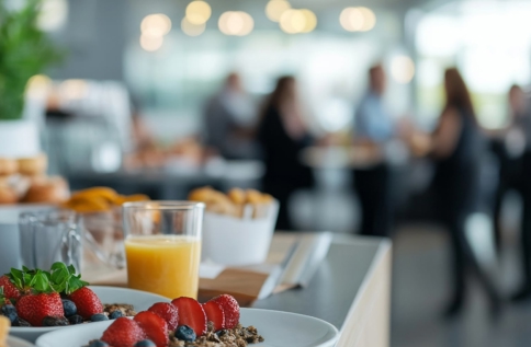 Frukostbuffé med jordgubbar, bröd och juice i förgrunden, människor samtalar suddigt i bakgrunden i ljus lokal.