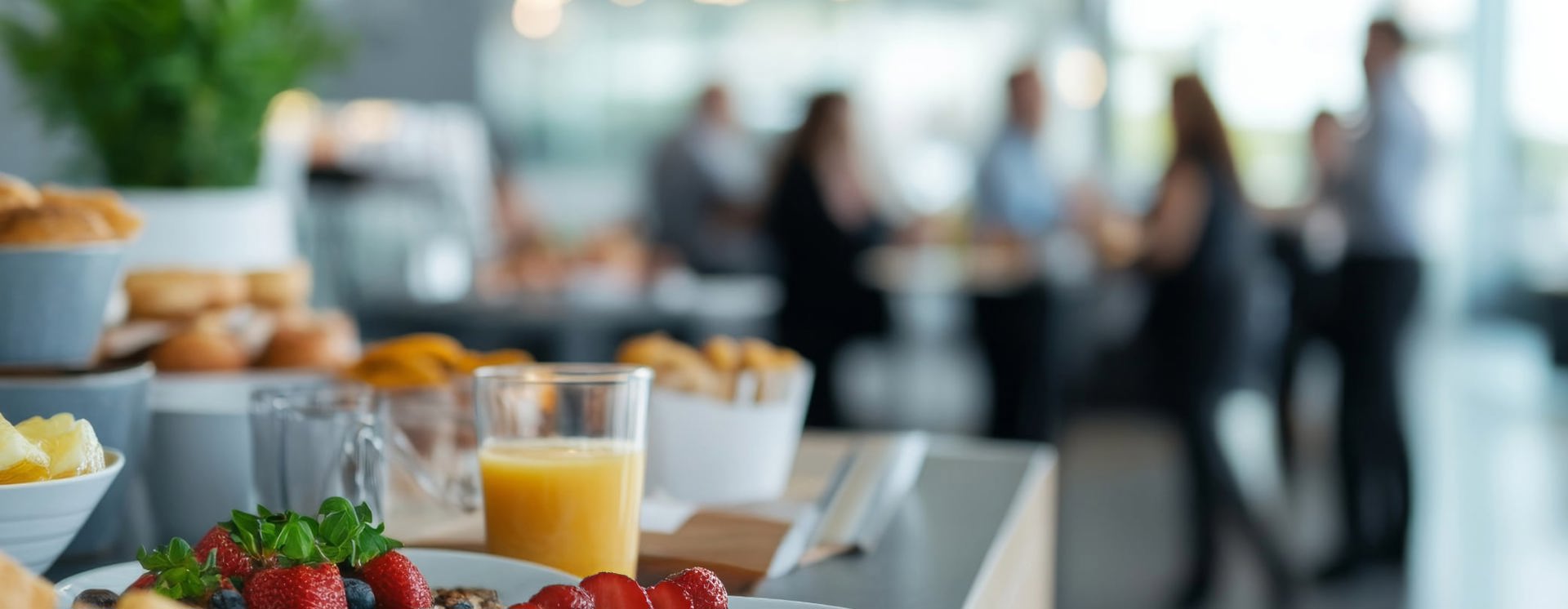 Frukostbuffé med jordgubbar, bröd och juice i förgrunden, människor samtalar suddigt i bakgrunden i ljus lokal.