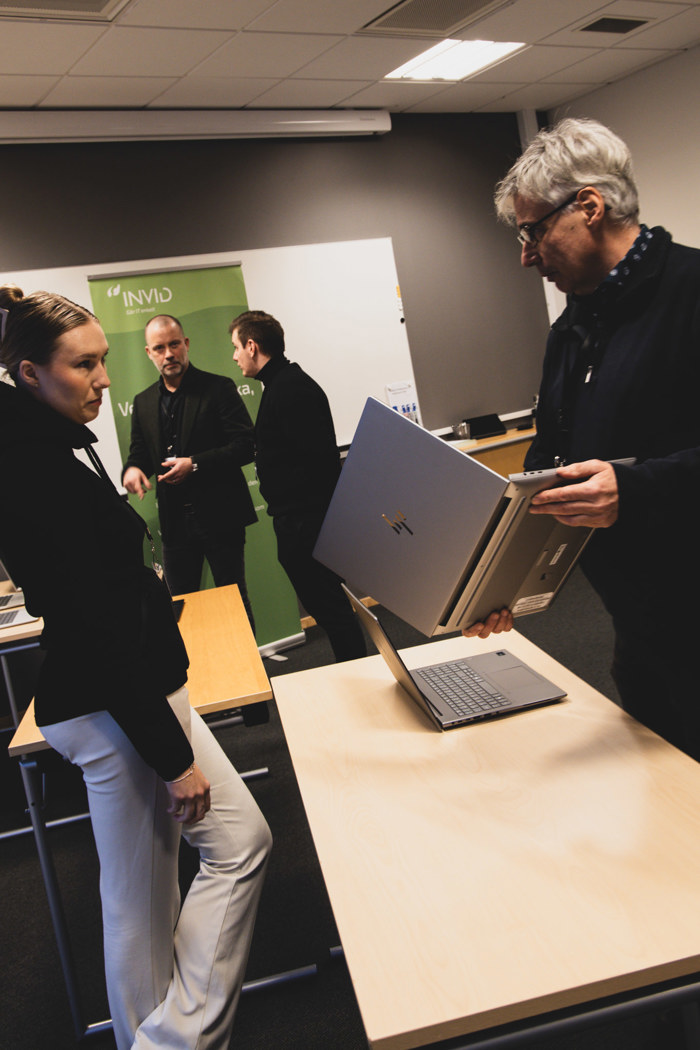 Person håller upp en laptop ovanför en annan på ett bord i ett konferensrum, medan andra personer samtalar i bakgrunden framför en INVID‑rollup