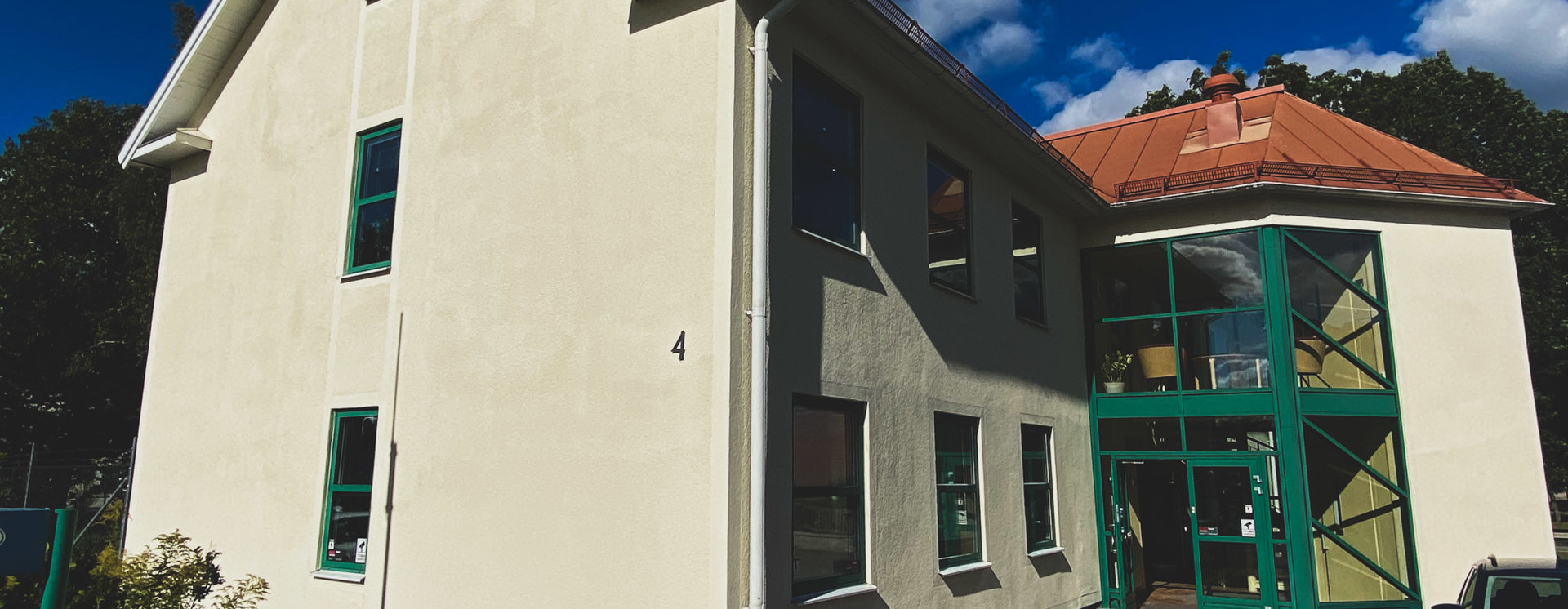 Ljus putsad byggnad i Anderstorp med grön glasentré och röda takdetaljer, sedd från utsidan under blå himmel, med gångväg och parkering i förgrunden