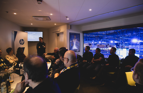 Personer sitter samlade i en loge och lyssnar på en presentation, med stor skärm och utsikt över upplyst arena i bakgrunden