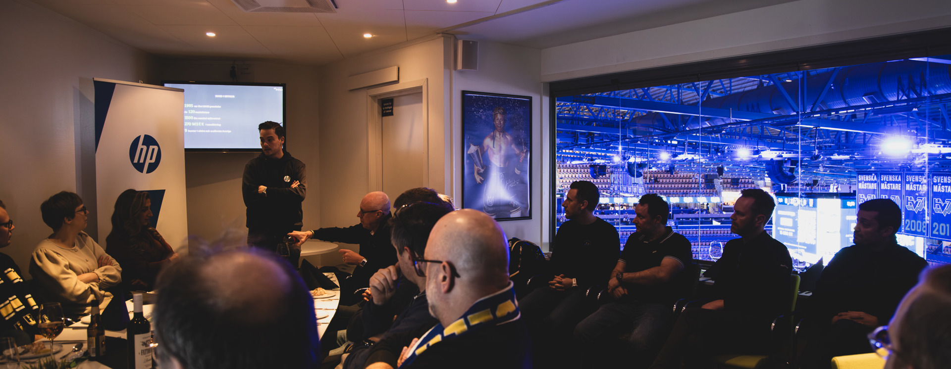 Personer sitter samlade i en loge och lyssnar på en presentation, med stor skärm och utsikt över upplyst arena i bakgrunden