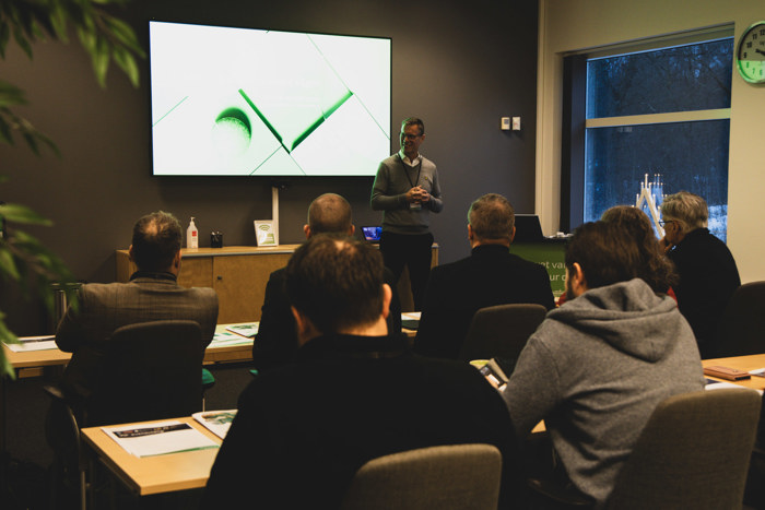 Johan Andersson presenterar framför en skärm i ett konferensrum medan deltagare sitter vid bord med anteckningar och material