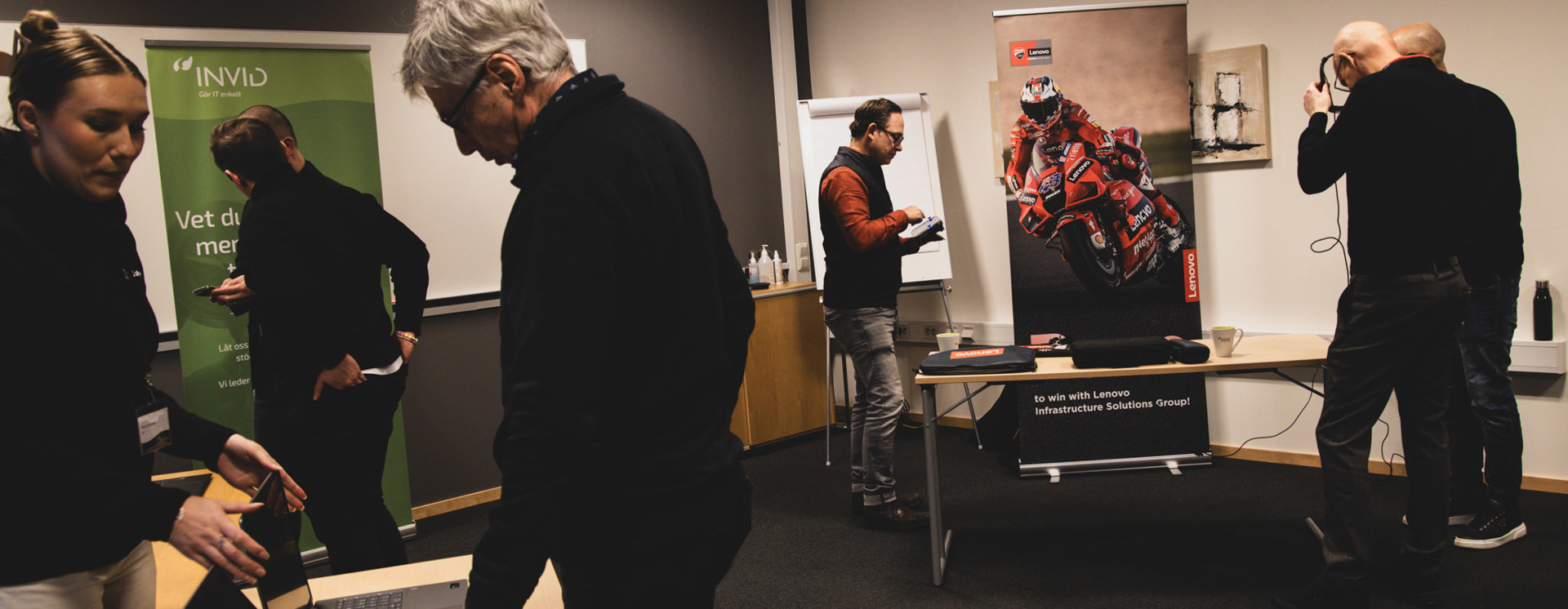 Flera personer rör sig i ett konferensrum kring bord med laptops, skärmar och material, med rollups och en racingbild i bakgrunden