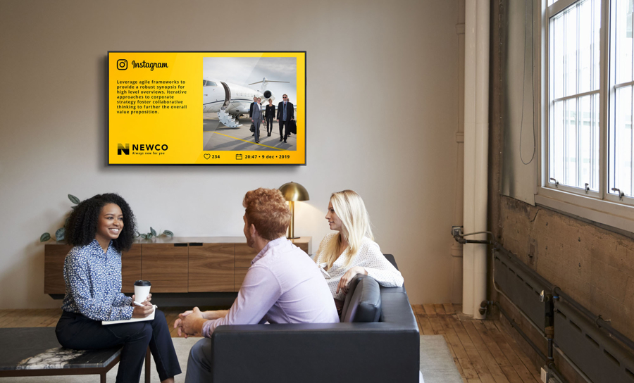 Tre personer sitter i lounge i kontorsmiljö och samtalar framför väggmonterad digital signage-skärm som visar marknadsinnehåll från sociala medier, vilket illustrerar intern kommunikation, varumärkesbyggande och informationsspridning