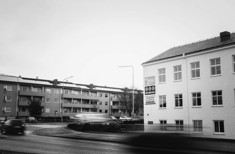 Svartvit bild på en vit byggnad i Västervik vid en rondell med tydlig INVID‑skylt på fasaden, omgiven av bostadshus och passerande trafik under en ljus himmel.