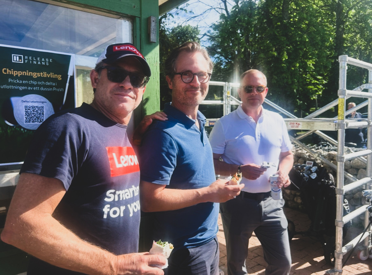 Tre deltagare samlas vid klubbhus under golftävling och äter lunch i solsken intill informationstavla och golfutrustning