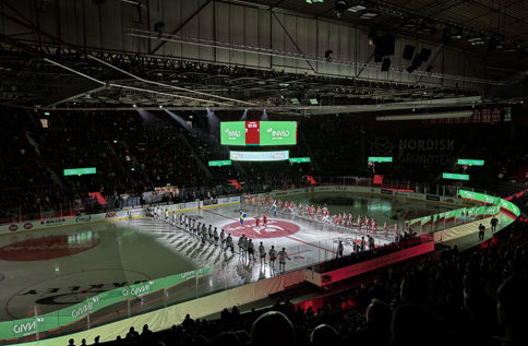 Hockeyarena med spelarpresentation på isen inför match, där INVID:s varumärke syns på skärmar och sargreklam framför stor publik