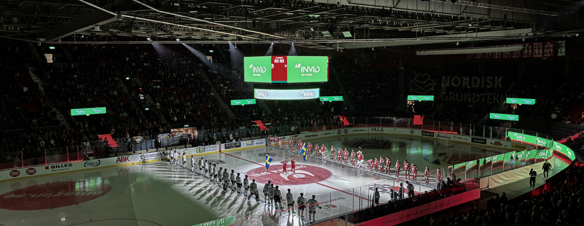 Hockeyarena med spelarpresentation på isen inför match, där INVID:s varumärke syns på skärmar och sargreklam framför stor publik