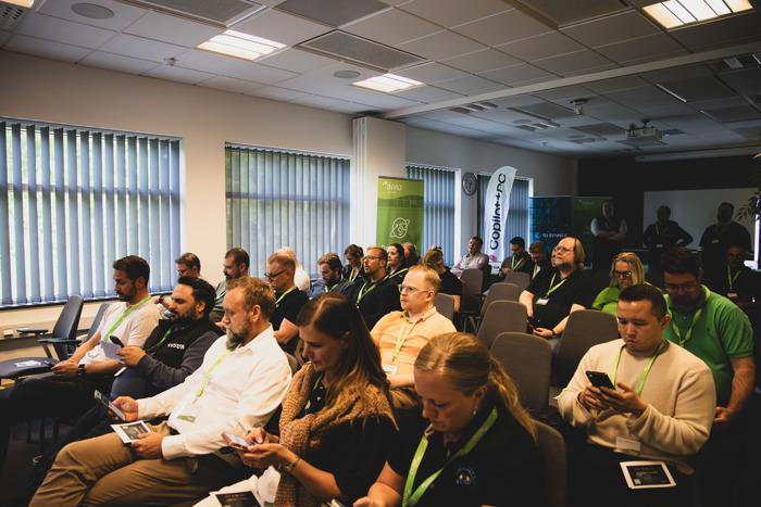Publik sitter i konferensrum under event och tittar på sina mobiltelefoner, med INVID‑ och Copilot+ PC‑flaggor synliga i bakgrunden.