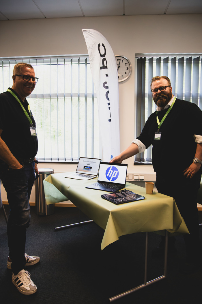 Två personer står vid ett demobord med laptops och informationsmaterial under event, med roll‑up i bakgrunden.