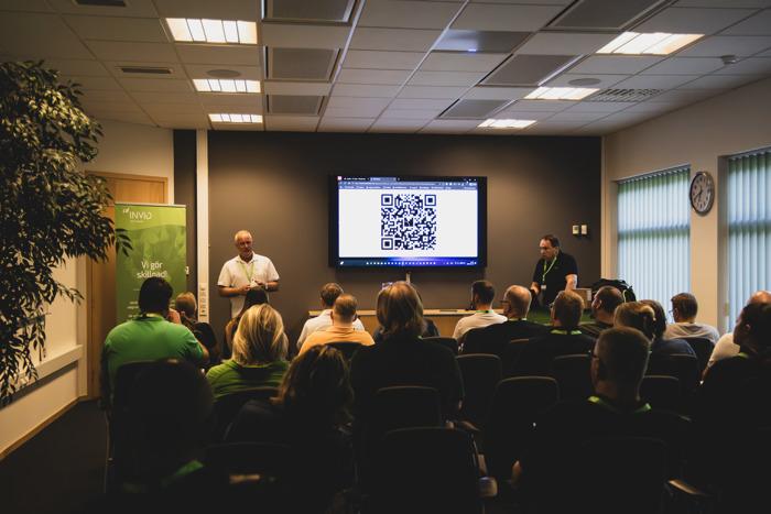 Föreläsare presenterar inför sittande publik i konferensrum, med QR‑kod visad på skärm och INVID‑banner vid sidan.
