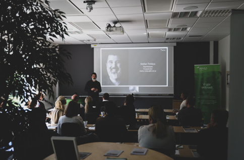 Stefan Timbus står och presenterar framför projicerad bild med porträtt och text i ett konferensrum, medan deltagare sitter vid bord och lyssnar
