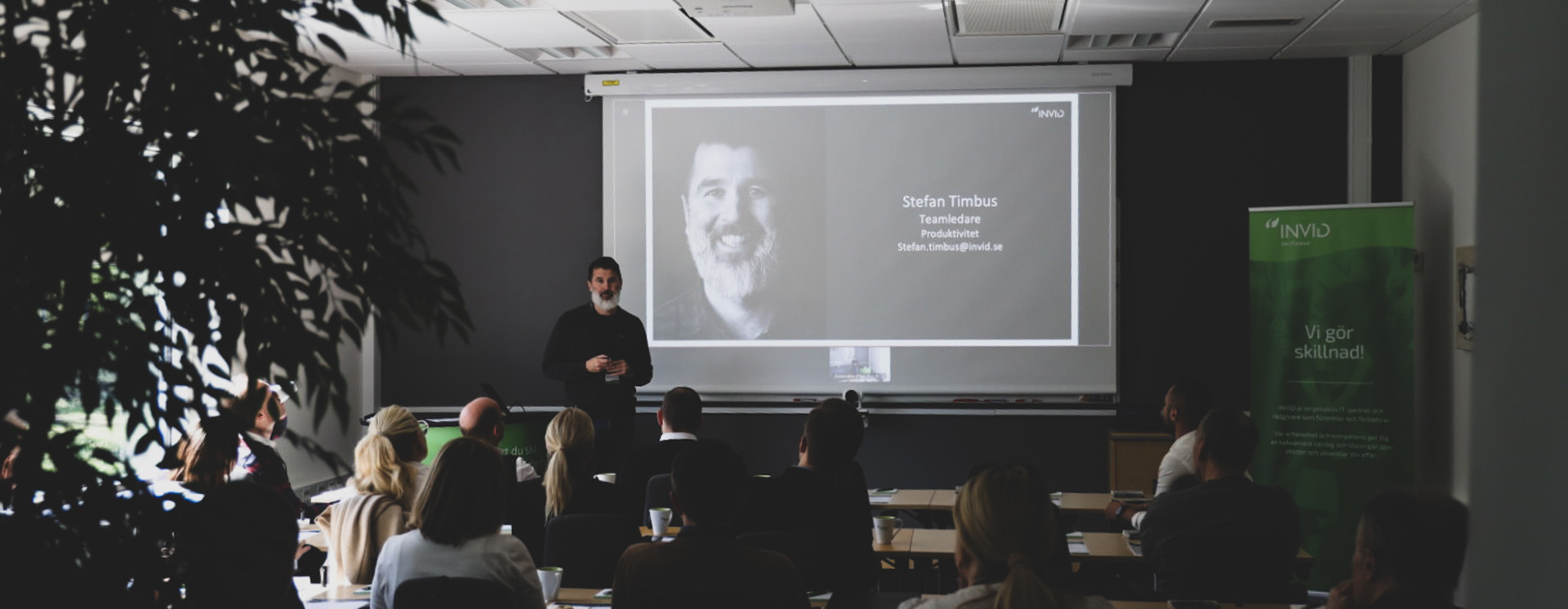 Stefan Timbus står och presenterar framför projicerad bild med porträtt och text i ett konferensrum, medan deltagare sitter vid bord och lyssnar