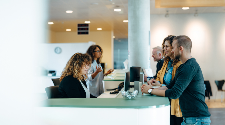 Personer vid reception i modern kontorsmiljö där besökare får hjälp av personal vid disk, med fokus på service, kommunikation och professionellt mottagande
