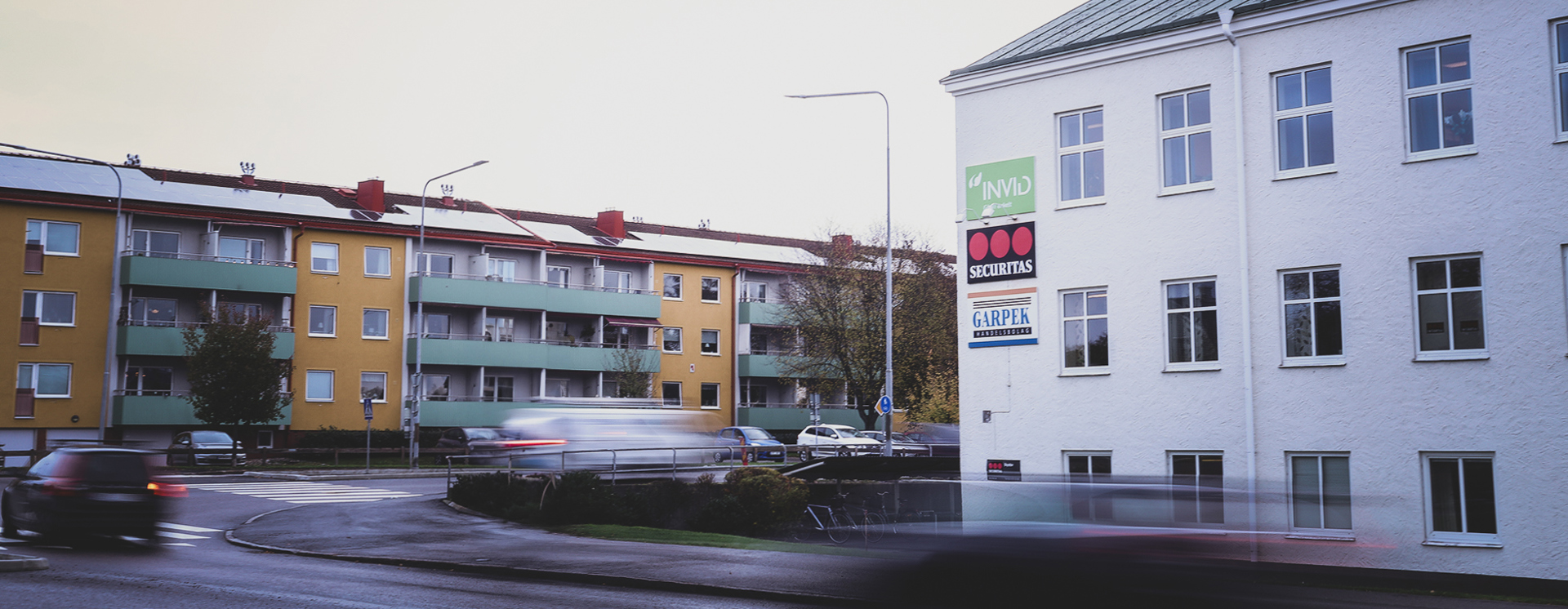 Vit byggnad i Västervik vid en rondell med tydlig INVID‑skylt på fasaden, omgiven av bostadshus och passerande trafik under en ljus himmel.