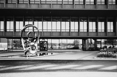 Svartvit bild på Viared Center i Borås, en modern byggnad med träfasad och stora glaspartier, med en reflekterande skulptur placerad vid en vattenyta framför entrén