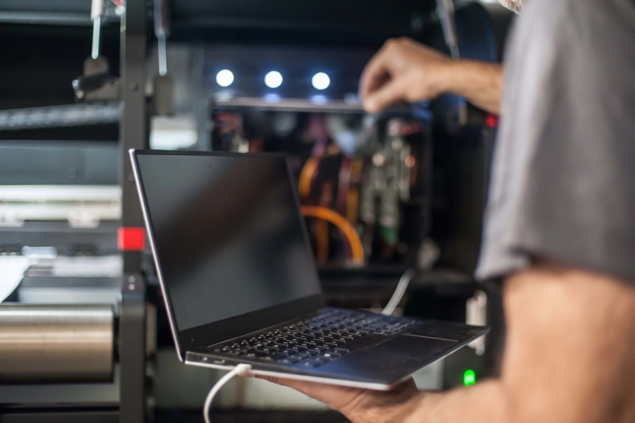 Person arbetar med laptop vid industriell maskin och utför tekniskt underhåll, vilket illustrerar IT‑stöd i produktion, felsökning och integration mellan system och maskiner i verksamheten