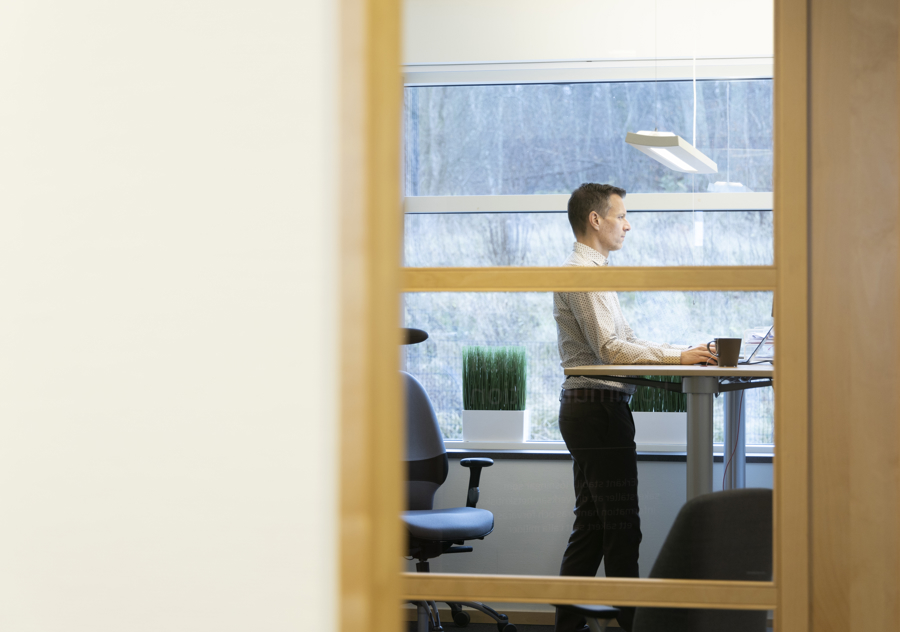 Person står vid höj- och sänkbart skrivbord i modern kontorsmiljö med laptop och kaffekopp, vilket visar ergonomisk arbetsplats, digitalt arbete och flexibel IT‑arbetsmiljö