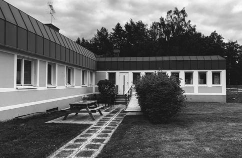 Svartvit bild på enplansbyggnad i Tranås med flera fönster och mörkt tak, sedd från en gräsmatta med gångstig, uteplats, bänk och buskar i förgrunden, samt träd och molnig himmel i bakgrunden