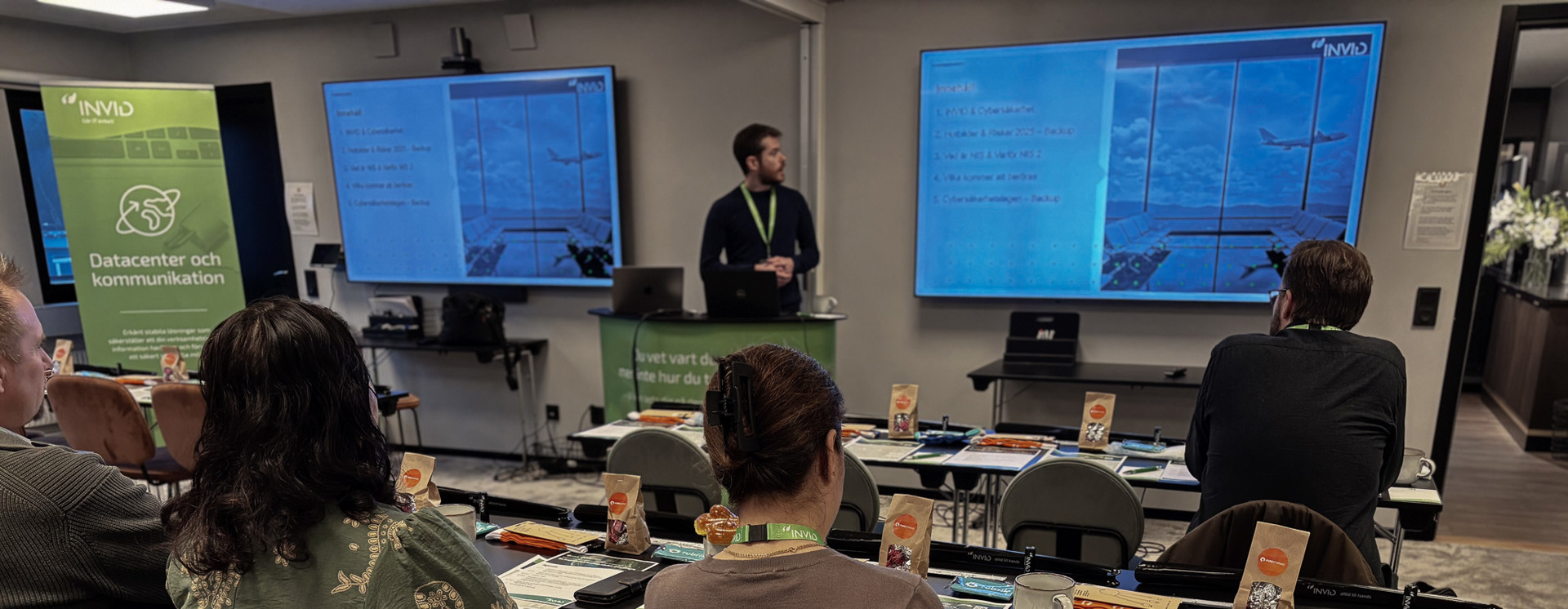 Person står och presenterar framför dubbla skärmar med presentation, medan deltagare sitter vid uppdukade bord och lyssnar i ett konferensrum