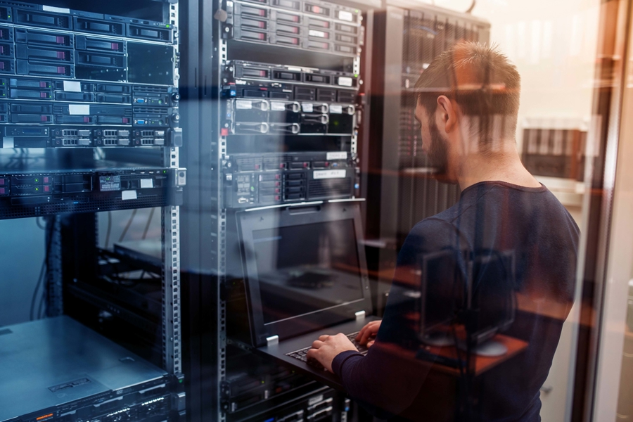 Tekniker arbetar vid laptop i serverhall framför rackmonterade servrar, vilket illustrerar serverdrift som tjänst, övervakning, felsökning och hantering av IT‑infrastruktur i modern datacentermiljö
