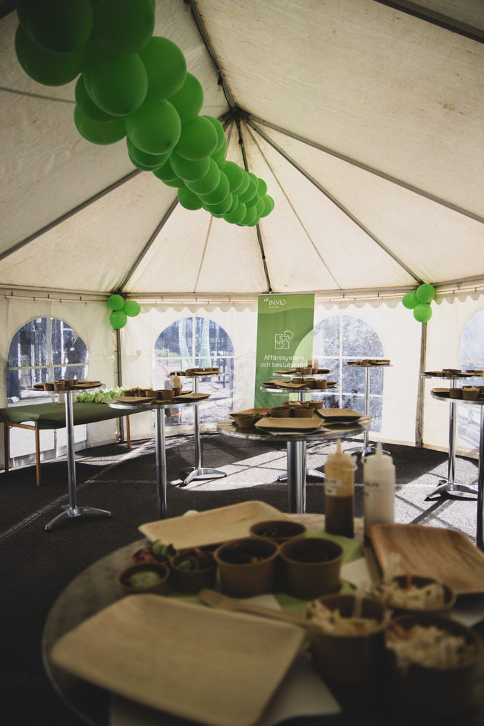 Serveringsyta i tält med ståbord och färdigställda matportioner, dekorerat med gröna ballonger och INVID‑skylt för affärssystem och beslutsstöd.